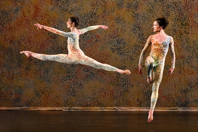 Two women in unitards with tiny colorful dots jump in front of a back drop with same dot pattern which is reminiscent of pointillism 